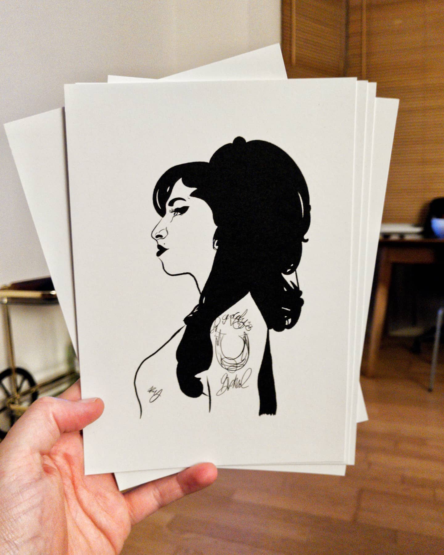 a photograph of a hand holding a small stack of paper to the camera, the front piece has an expressive black llustration of Amy Winehouse